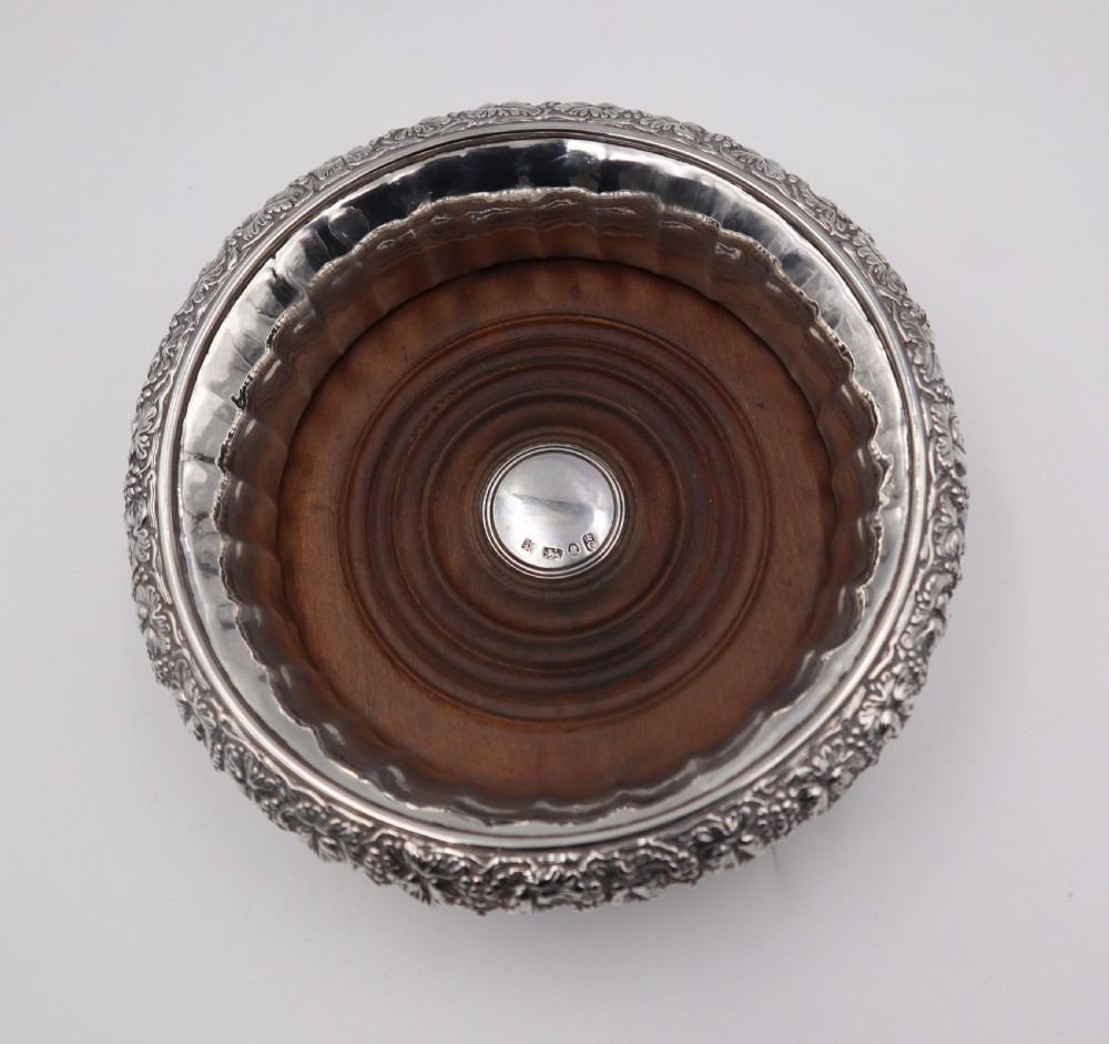 A set of four circular silver wine coasters featuring fluted inner rims, embossed grape and vine borders, and turned wood bases with central silver bosses. View V