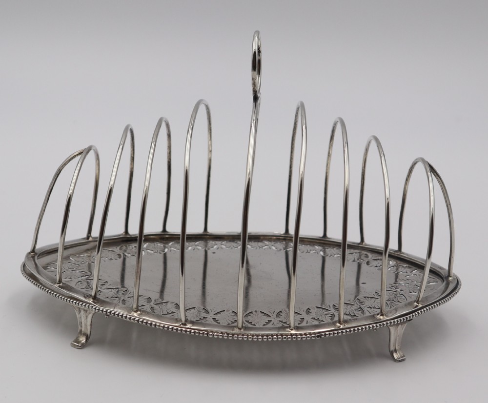 An oval antique silver toast rack featuring graduated wire arches, a loop handle, an engraved floral base border, and small feet. View II