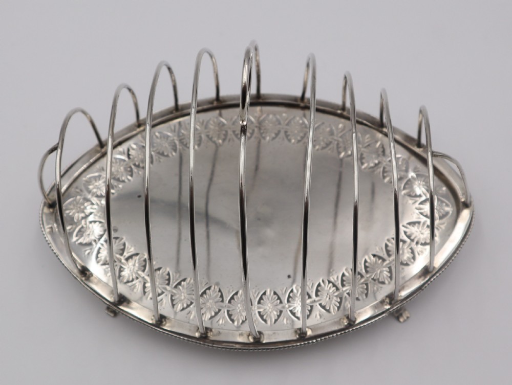 An oval antique silver toast rack featuring graduated wire arches, a loop handle, an engraved floral base border, and small feet. View III