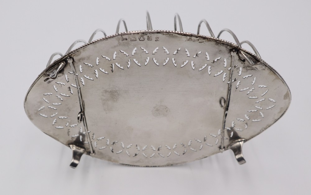An oval antique silver toast rack featuring graduated wire arches, a loop handle, an engraved floral base border, and small feet. View IV