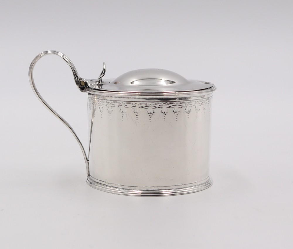 A cylindrical sterling silver mustard pot featuring a domed hinged lid with a thumbpiece, a loop handle, and engraved bright-cut detailing around the upper rim.