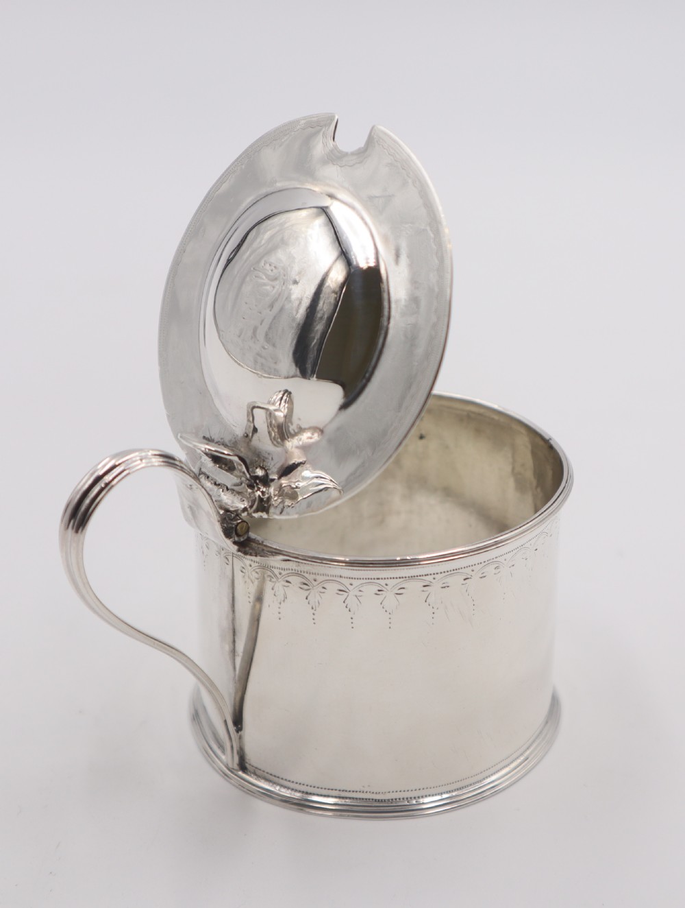 A cylindrical sterling silver mustard pot featuring a domed hinged lid with a thumbpiece, a loop handle, and engraved bright-cut detailing around the upper rim. View V