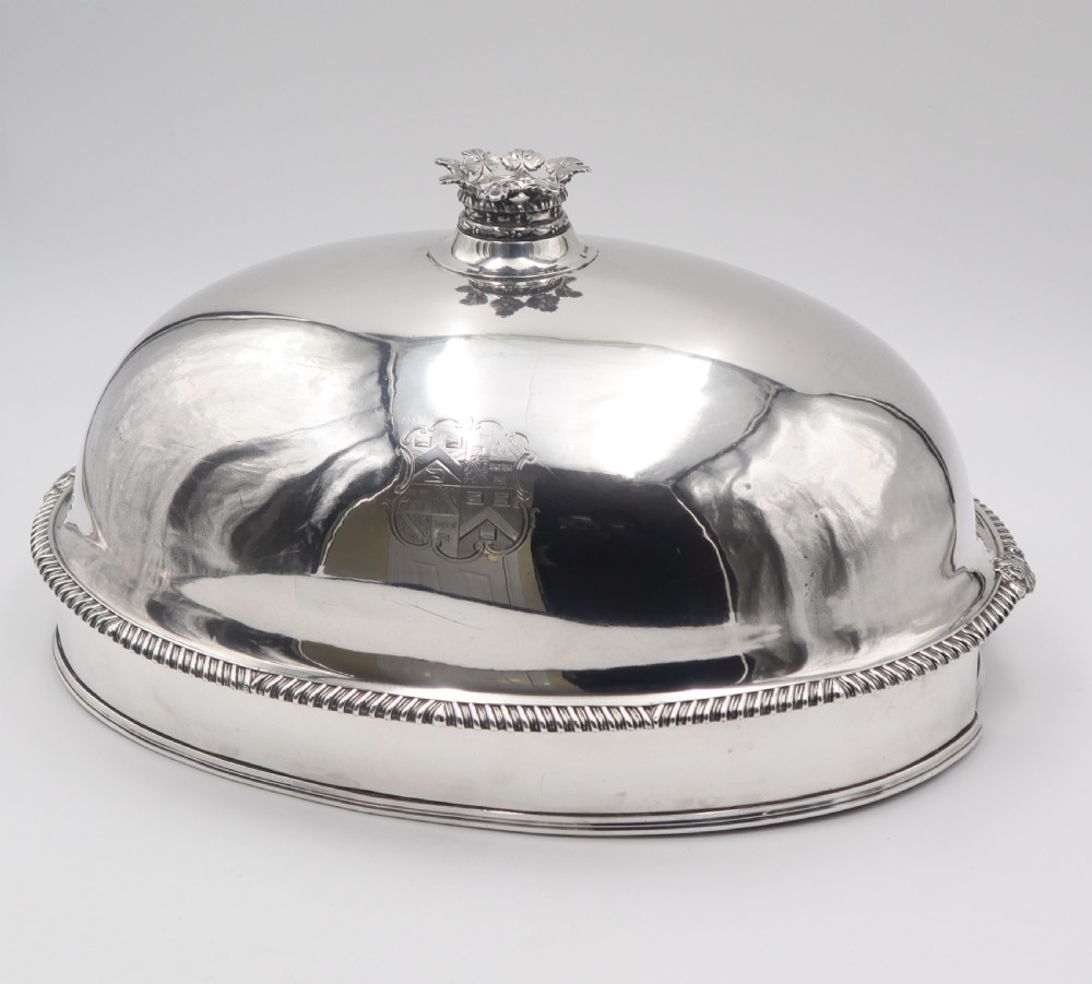 An oval silver-plated meat dome featuring a gadrooned rim, an ornate foliate handle, and an engraved coat of arms on the polished body.