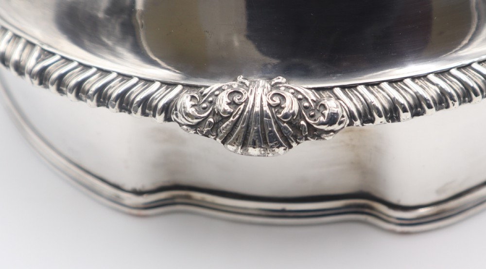 An oval silver-plated meat dome featuring a gadrooned rim, an ornate foliate handle, and an engraved coat of arms on the polished body. View V