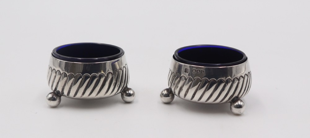 A pair of circular silver salt cellars featuring spiral-fluted bodies, three small ball feet, and removable cobalt blue glass liners.
