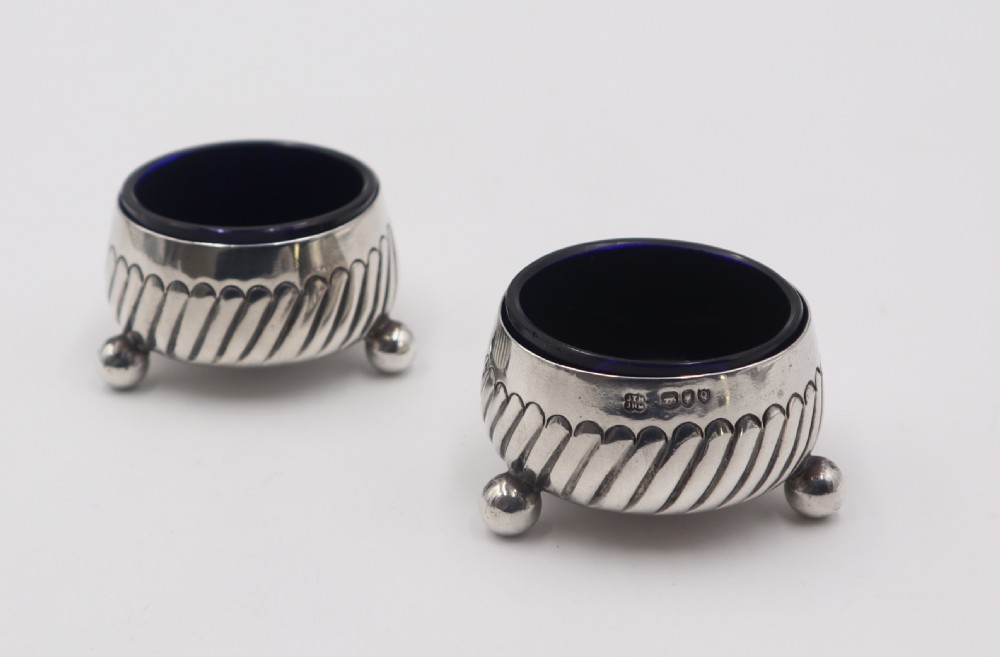 A pair of circular silver salt cellars featuring spiral-fluted bodies, three small ball feet, and removable cobalt blue glass liners. View II