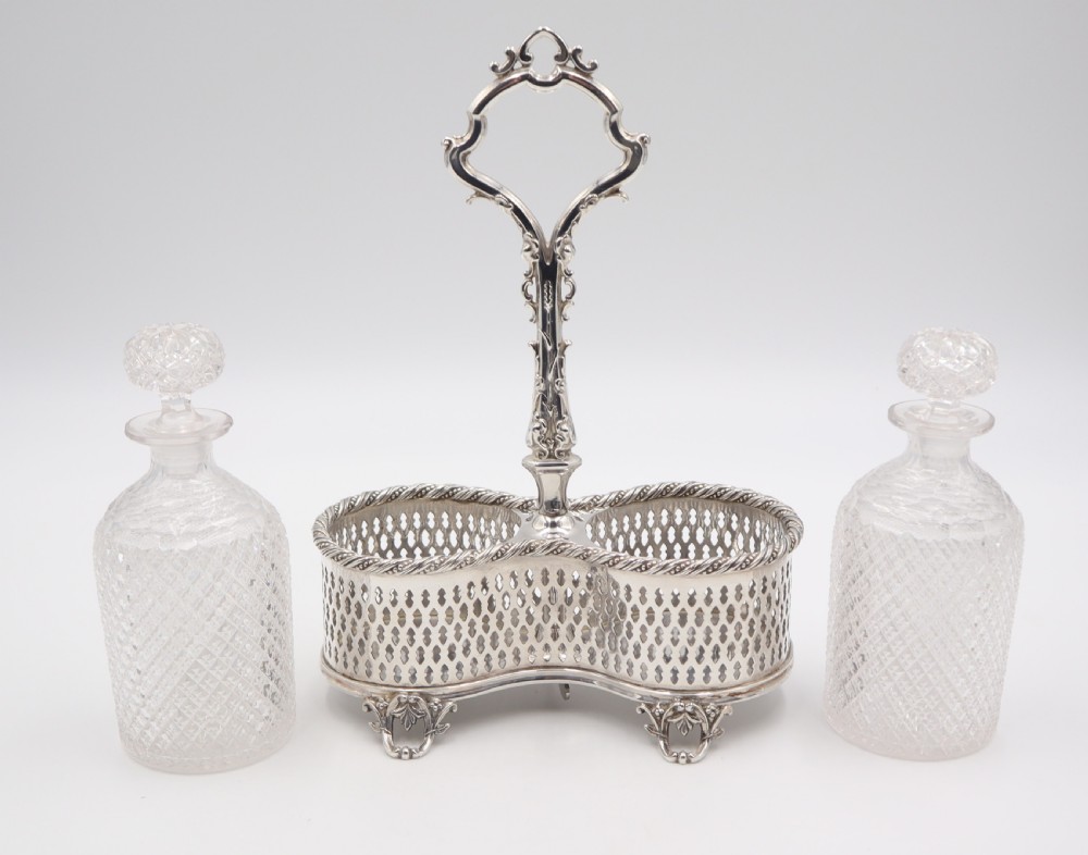 An antique silver-plated liqueur stand featuring a pierced lattice gallery, decorative central handle, and scroll feet, holding two cut-glass decanters with stoppers. View III
