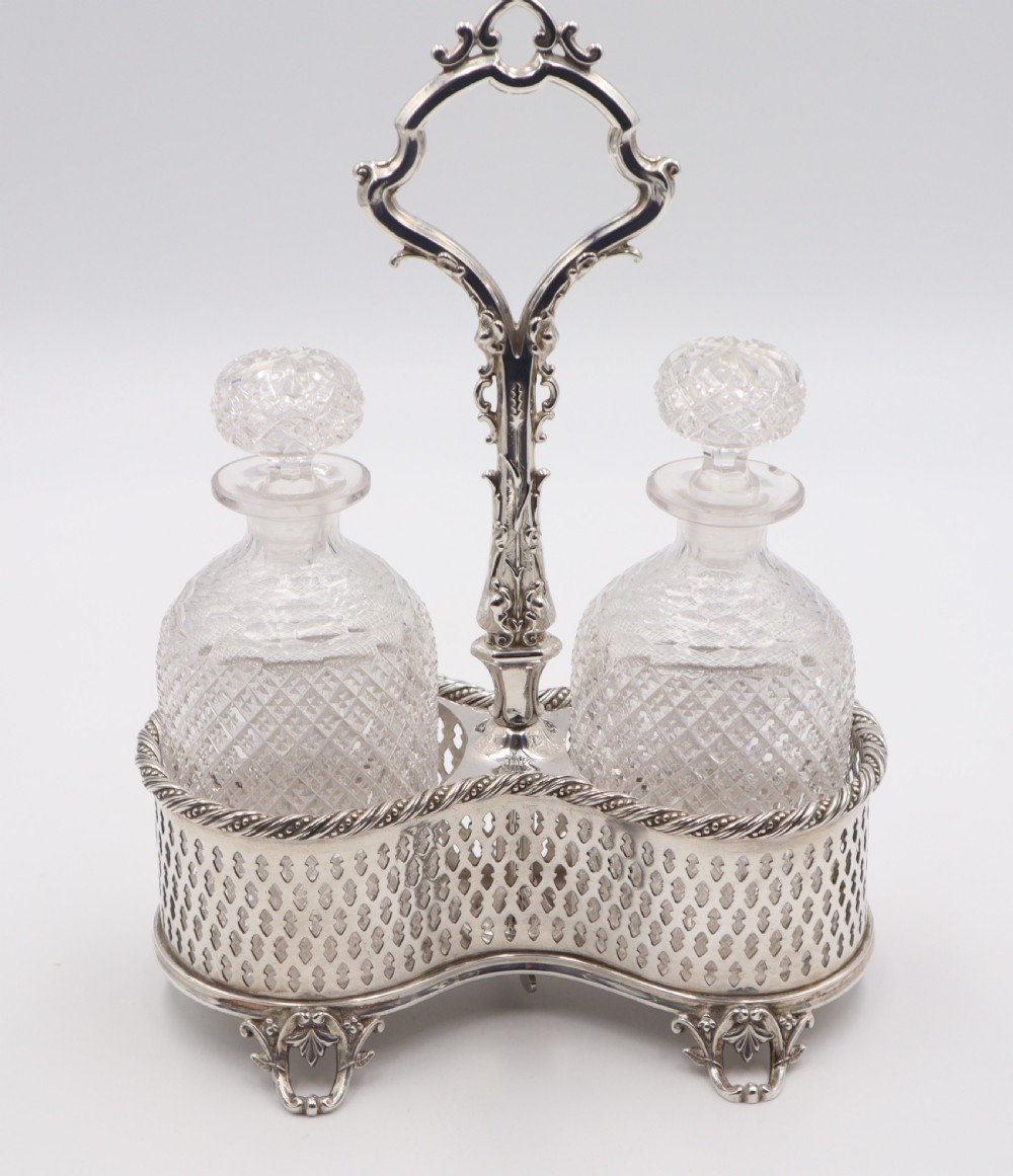 An antique silver-plated liqueur stand featuring a pierced lattice gallery, decorative central handle, and scroll feet, holding two cut-glass decanters with stoppers. View VIII