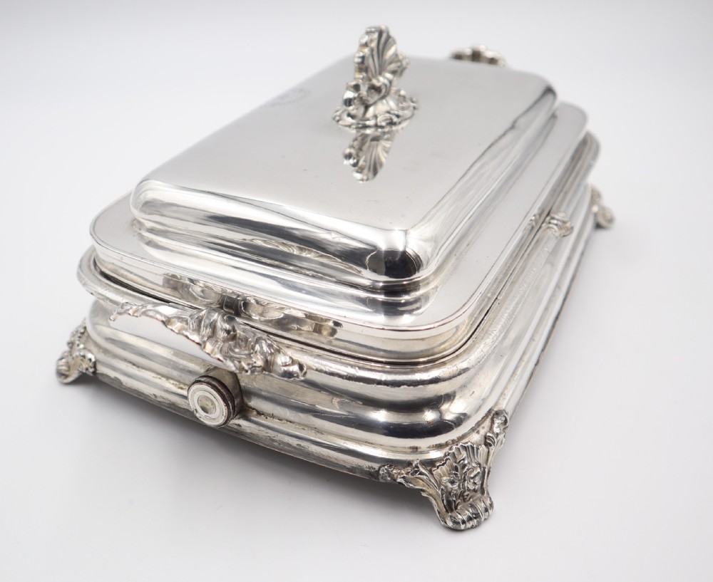 A rectangular silver-plated warming entree dish featuring shell-form handles, a domed lid with a shell finial, and a raised pedestal base. View V