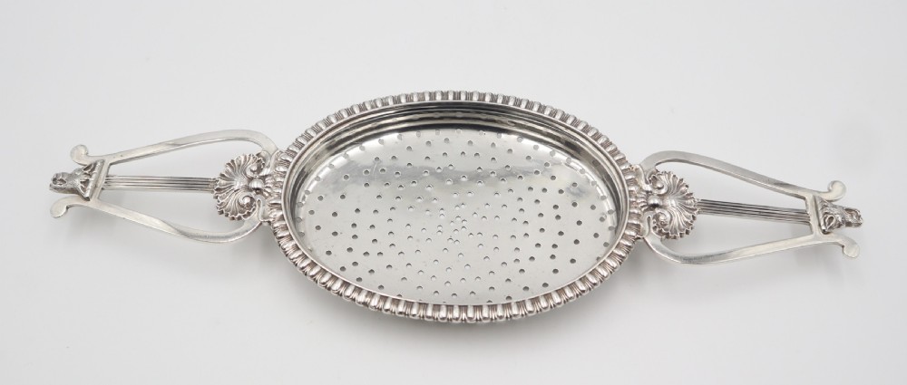 An antique oval sterling silver strainer featuring a perforated bowl, gadrooned rim, and two lyre-shaped handles adorned with shell details.