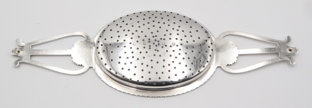 An antique oval sterling silver strainer featuring a perforated bowl, gadrooned rim, and two lyre-shaped handles adorned with shell details. View II