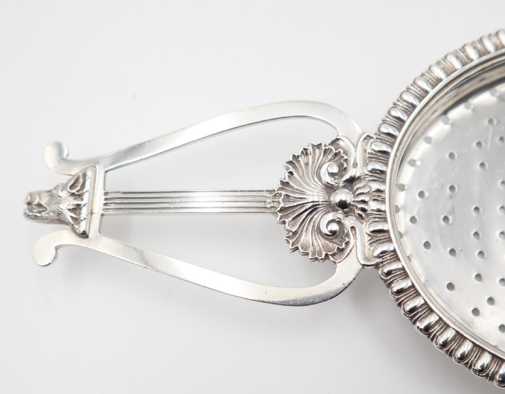 An antique oval sterling silver strainer featuring a perforated bowl, gadrooned rim, and two lyre-shaped handles adorned with shell details. View III