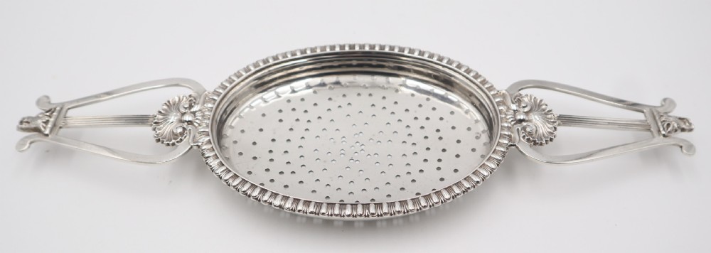 An antique oval sterling silver strainer featuring a perforated bowl, gadrooned rim, and two lyre-shaped handles adorned with shell details. View V