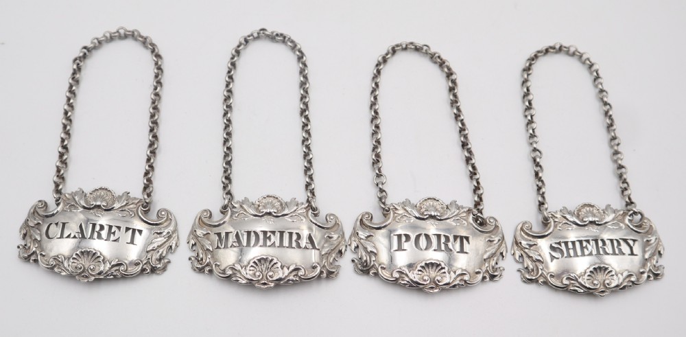 A set of four silver decanter labels featuring shell and scroll borders, each suspended by a chain and inscribed with Claret, Madeira, Port, and Sherry.