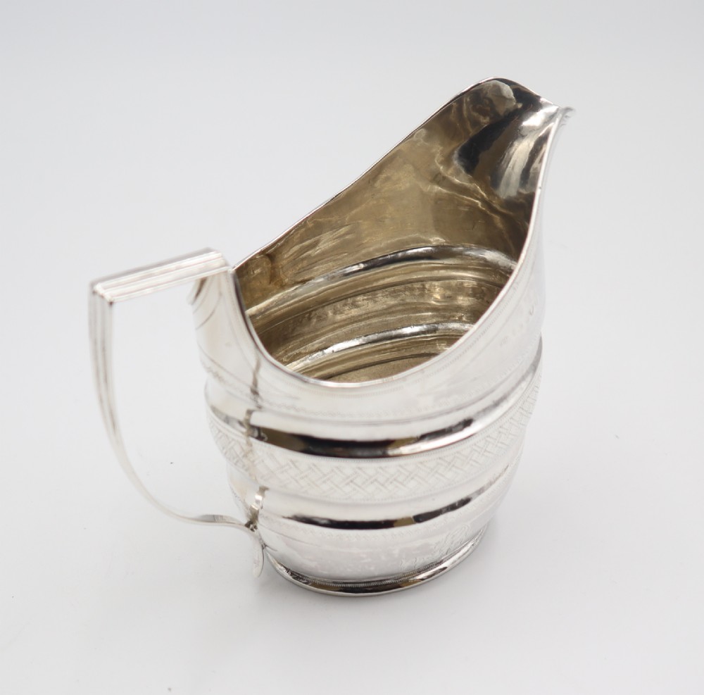 An antique sterling silver cream jug featuring geometric engraved banding, reeded body contours, an angular strap handle, and a polished metallic surface. View V
