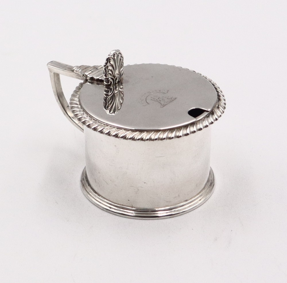 A cylindrical silver mustard pot with a hinged flat lid, gadrooned rim border, shell thumbpiece, angular handle, and an engraved crest.