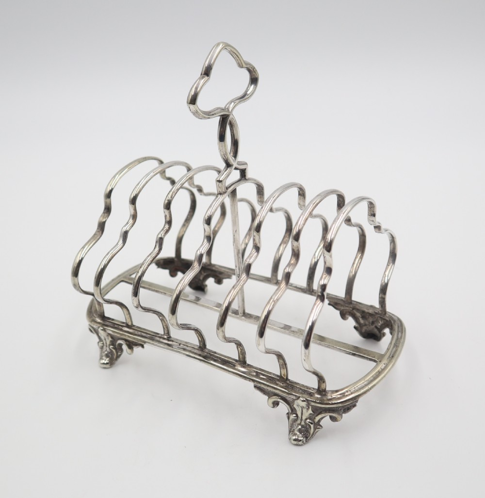 An antique silver plated toast rack featuring shaped wire dividers, a central loop handle, and cast scroll feet.