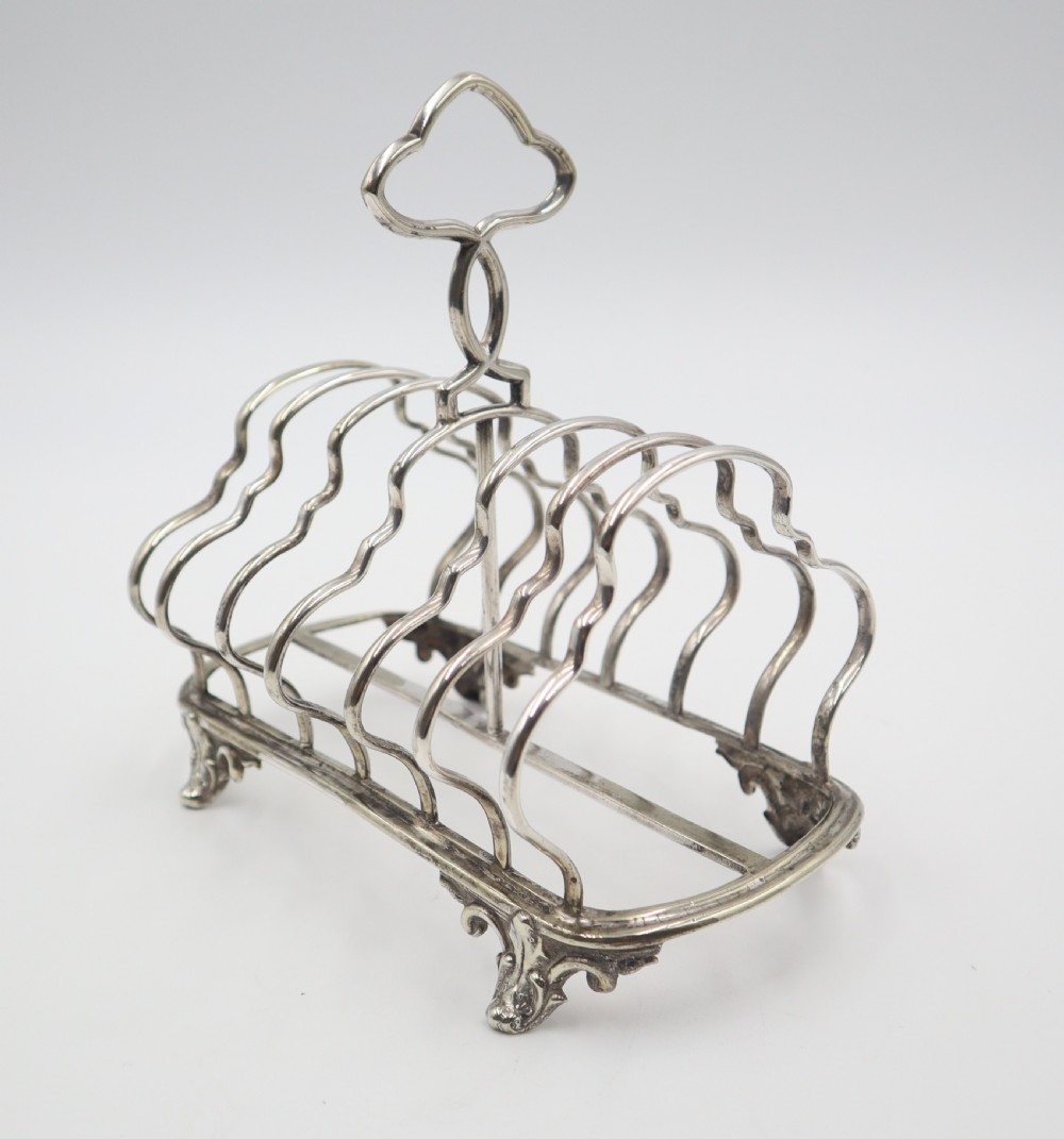 An antique silver plated toast rack featuring shaped wire dividers, a central loop handle, and cast scroll feet. View II