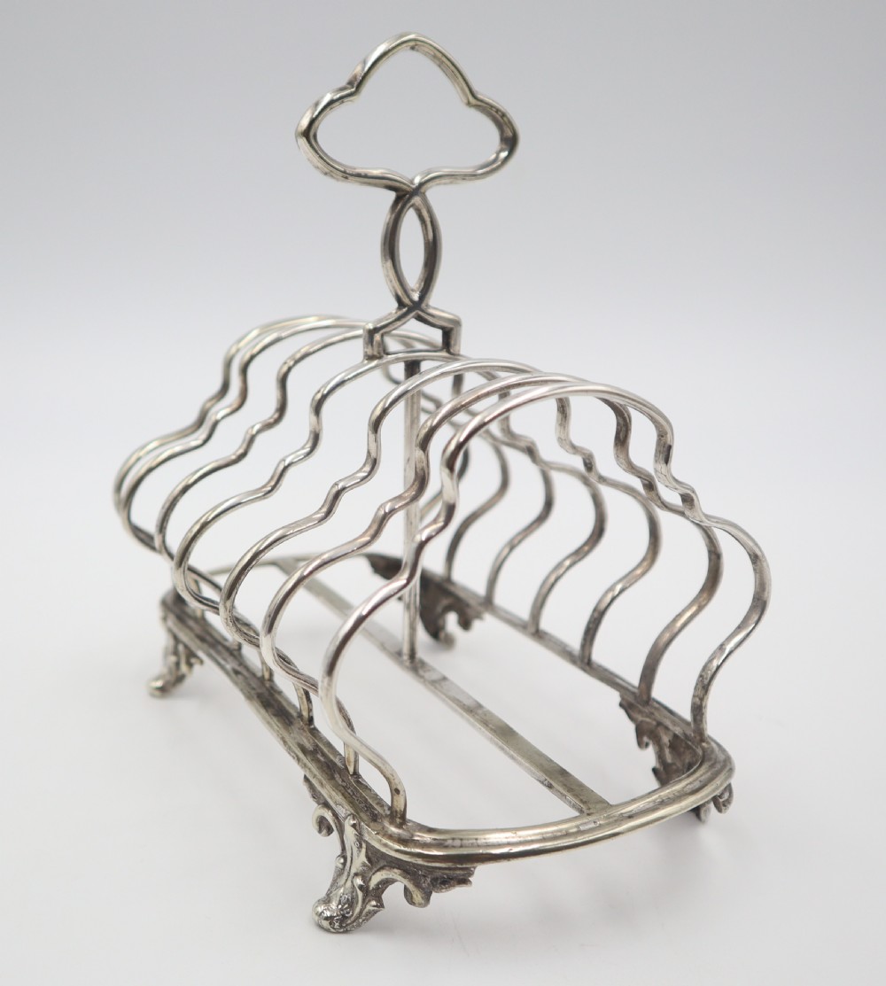 An antique silver plated toast rack featuring shaped wire dividers, a central loop handle, and cast scroll feet. View IV