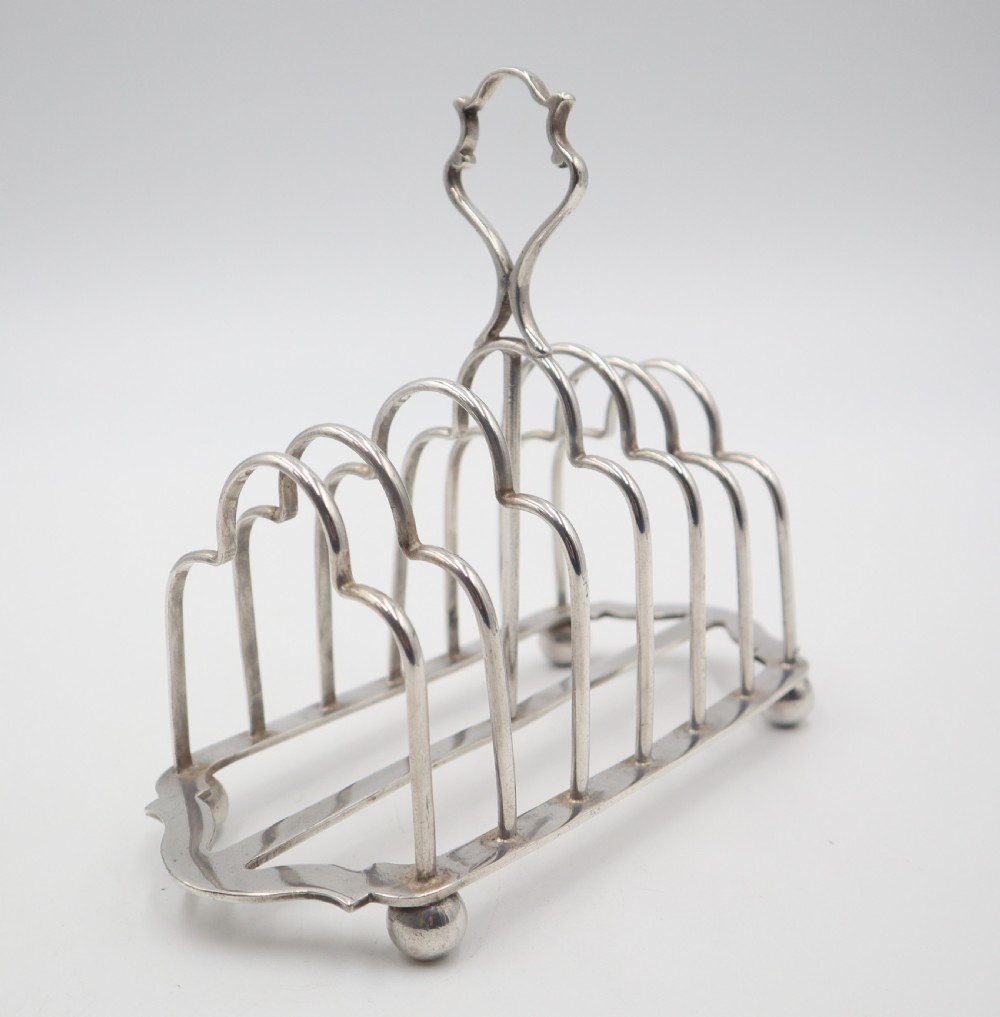 A silverplated toast rack featuring six arched wire slots, a central loop handle, and four small ball feet. View IV