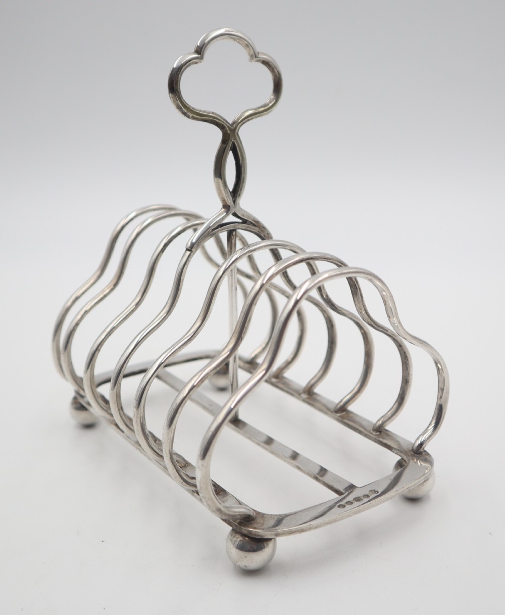 A silver-plated toast rack featuring six curved wire dividers, a central trefoil carrying handle, and four spherical ball feet.