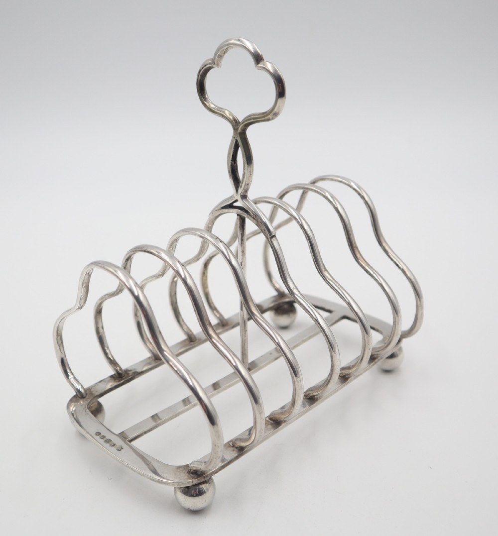 A silver-plated toast rack featuring six curved wire dividers, a central trefoil carrying handle, and four spherical ball feet. View IV