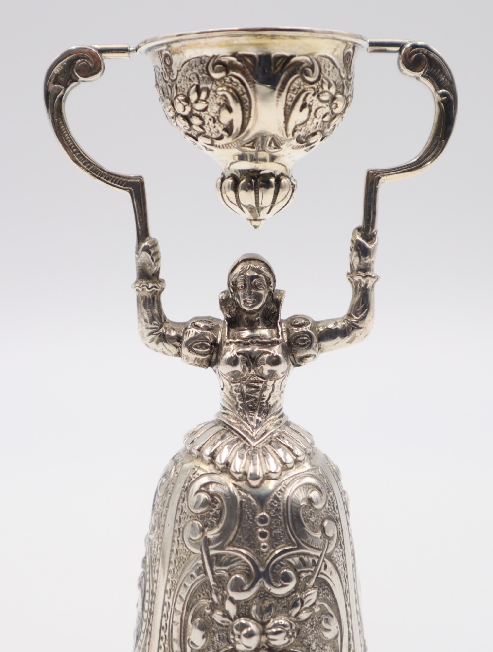 An antique silver wager cup shaped as a female figure in a detailed floral skirt holding an articulated secondary cup overhead. View V
