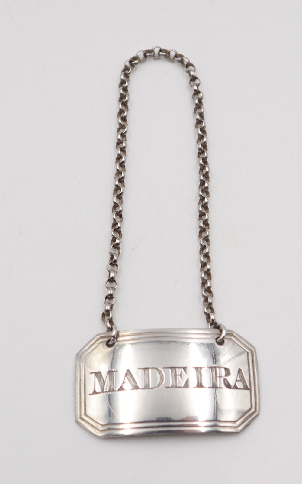 An antique rectangular sterling silver decanter label with canted corners engraved with the word MADEIRA and attached to a silver link suspension chain.