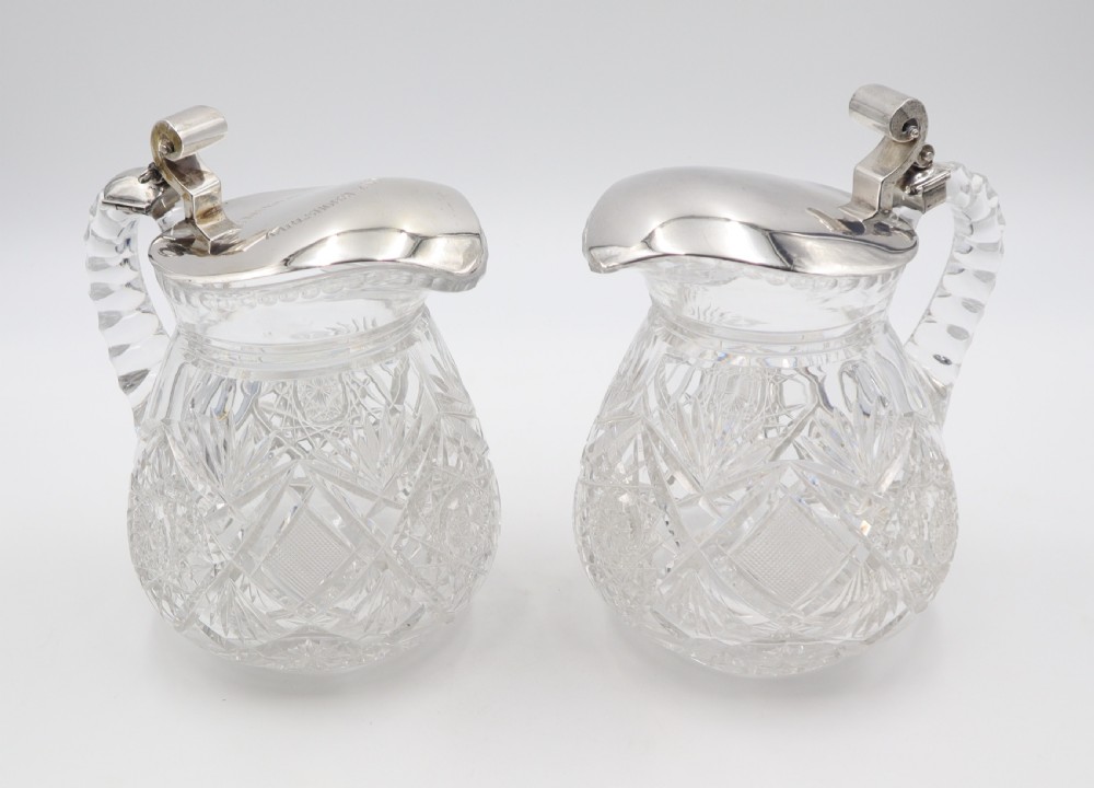 A pair of cut crystal glass jugs featuring silver mounts, hinged silver lids with scroll thumbpieces, and faceted crystal handles. View II