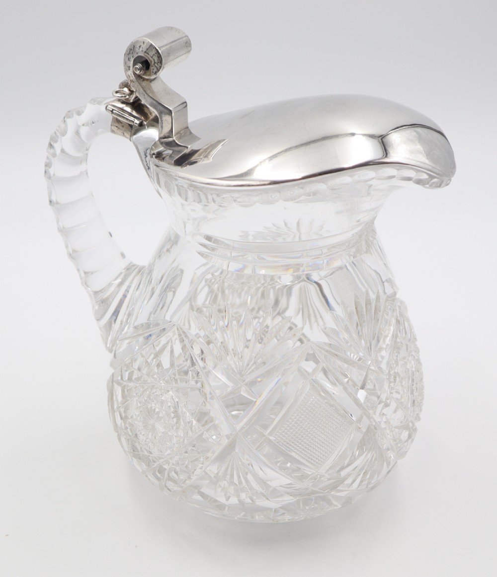 A pair of cut crystal glass jugs featuring silver mounts, hinged silver lids with scroll thumbpieces, and faceted crystal handles. View VII