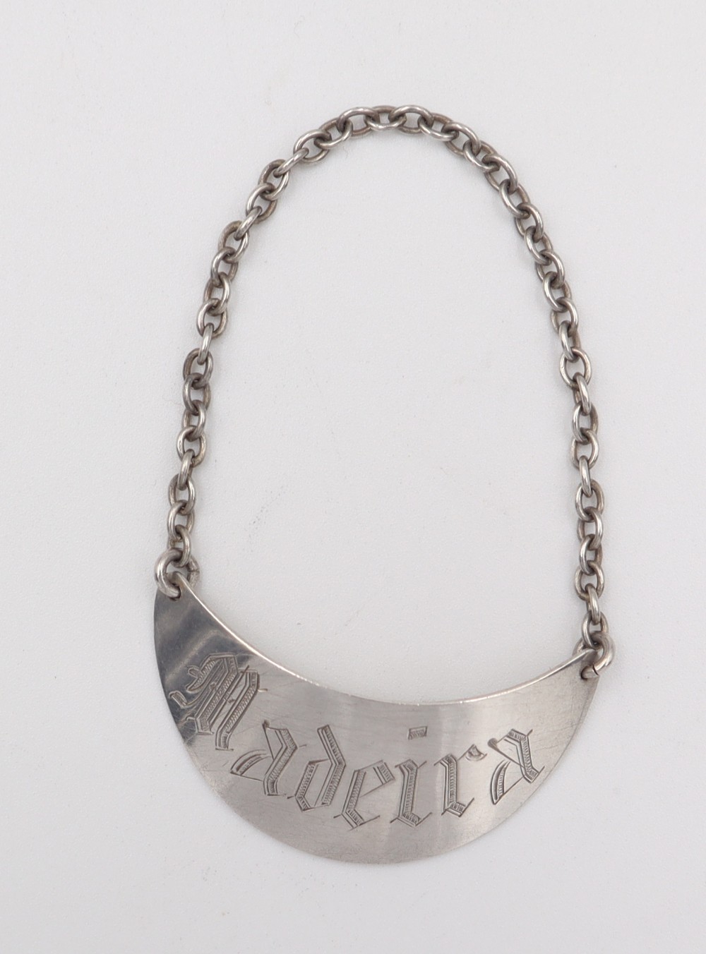 A crescent-shaped silver decanter label engraved with the word Madeira in Gothic script, attached to a silver suspension chain.