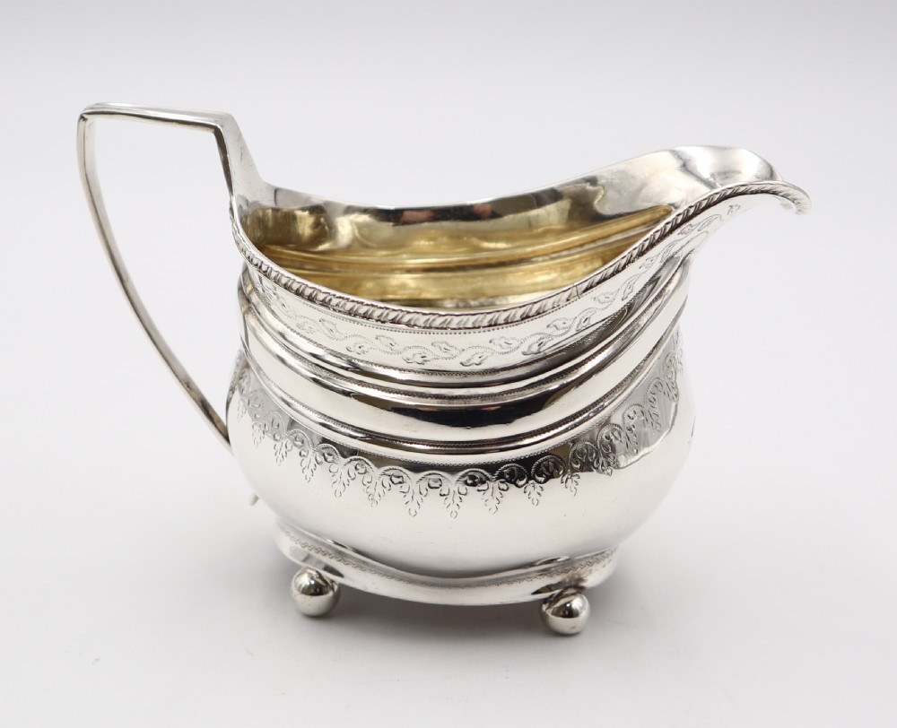 An antique sterling silver cream jug featuring engraved decorative borders, an angular handle, a gilded interior, and four small ball feet.
