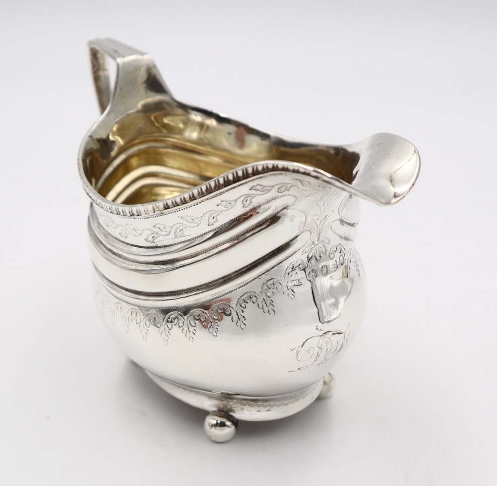 An antique sterling silver cream jug featuring engraved decorative borders, an angular handle, a gilded interior, and four small ball feet. View II