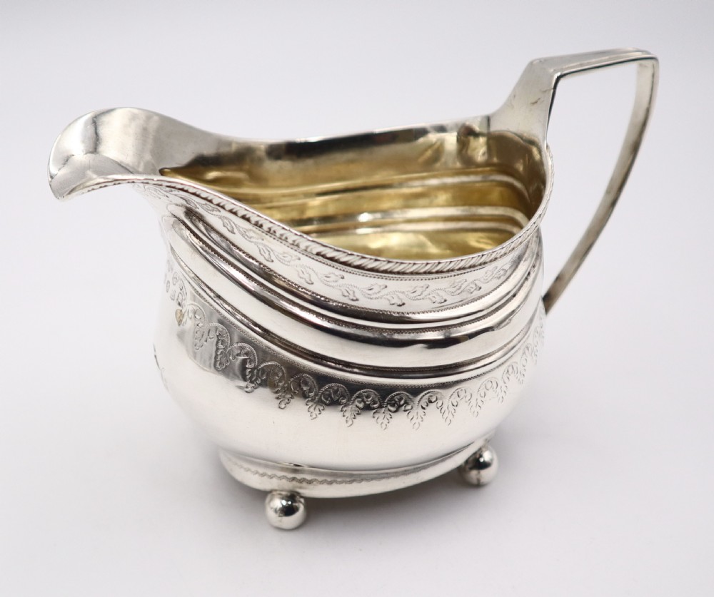 An antique sterling silver cream jug featuring engraved decorative borders, an angular handle, a gilded interior, and four small ball feet. View IV