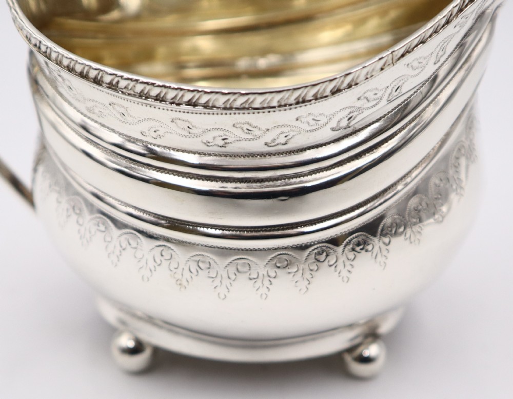 An antique sterling silver cream jug featuring engraved decorative borders, an angular handle, a gilded interior, and four small ball feet. View V