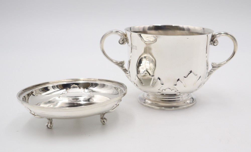 A two-handled sterling silver porringer cup featuring relief leaf decoration near the base beside a matching footed cover.