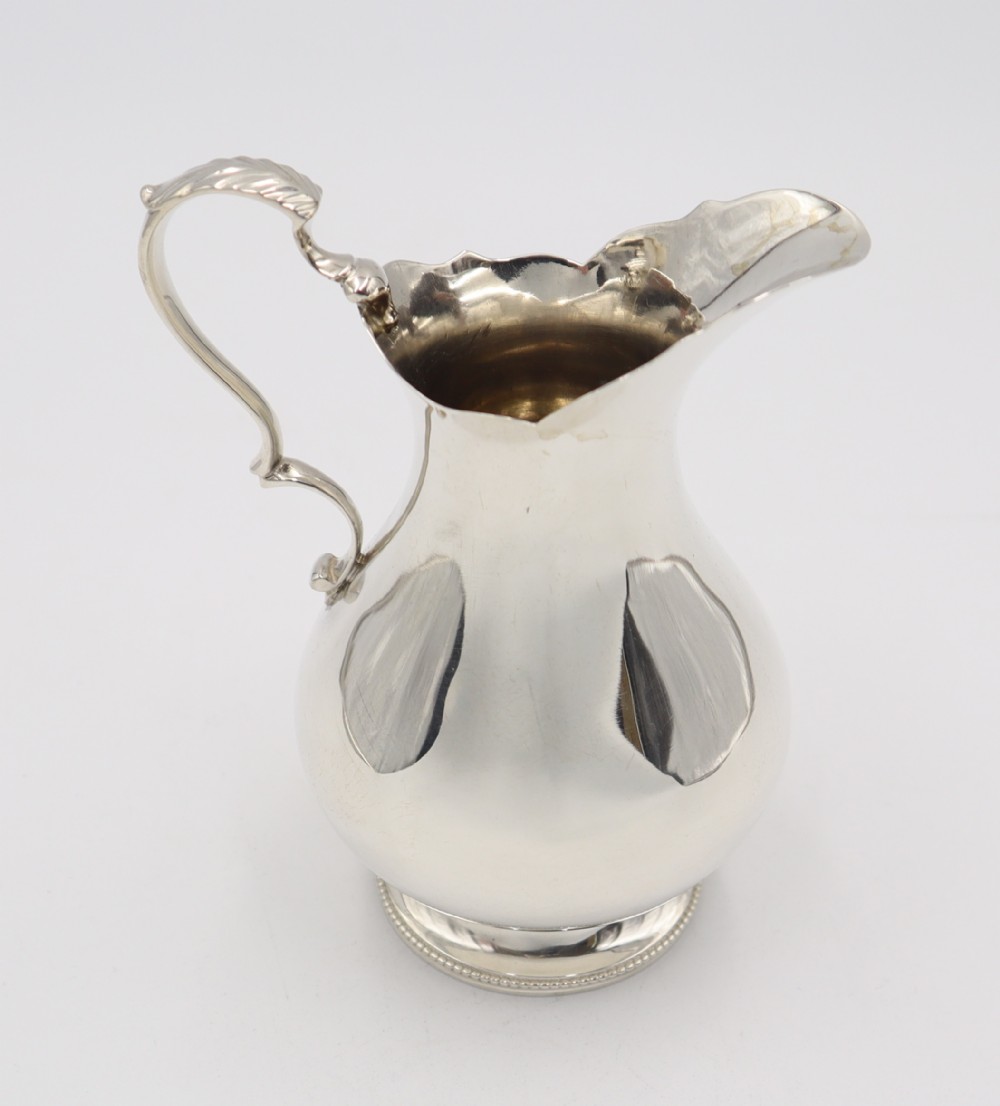 An antique silver cream jug featuring a baluster body, wavy scalloped rim, scroll handle with leaf detail, and a beaded circular base.