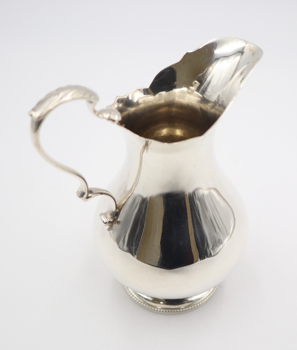 An antique silver cream jug featuring a baluster body, wavy scalloped rim, scroll handle with leaf detail, and a beaded circular base. View IV