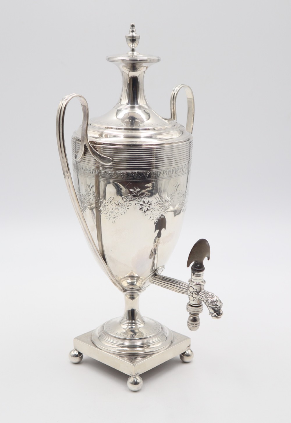An antique silver tea urn featuring high loop handles, engraved floral bands, a front tap, and a square pedestal base supported by ball feet.