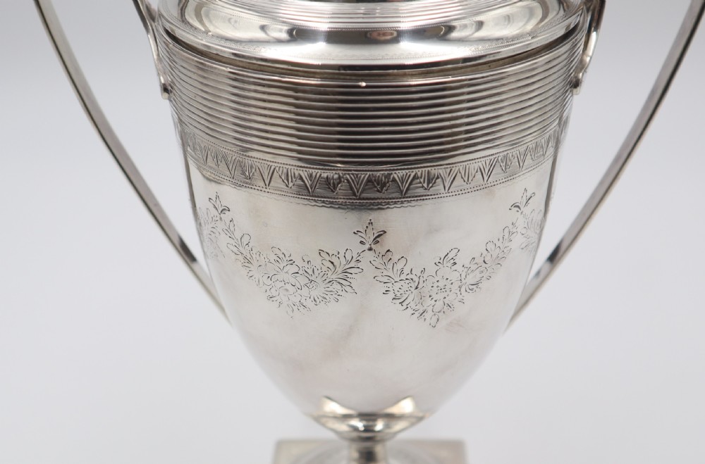 An antique silver tea urn featuring high loop handles, engraved floral bands, a front tap, and a square pedestal base supported by ball feet. View V