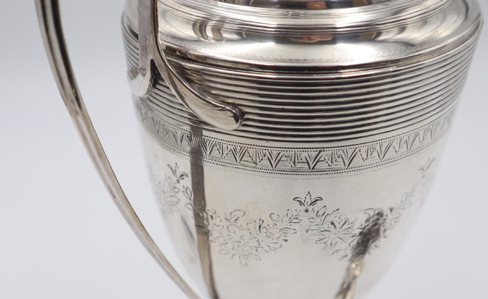 An antique silver tea urn featuring high loop handles, engraved floral bands, a front tap, and a square pedestal base supported by ball feet. View VI