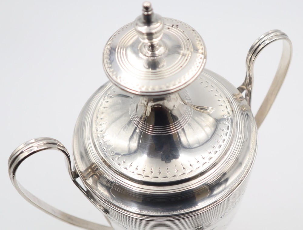 An antique silver tea urn featuring high loop handles, engraved floral bands, a front tap, and a square pedestal base supported by ball feet. View VIII