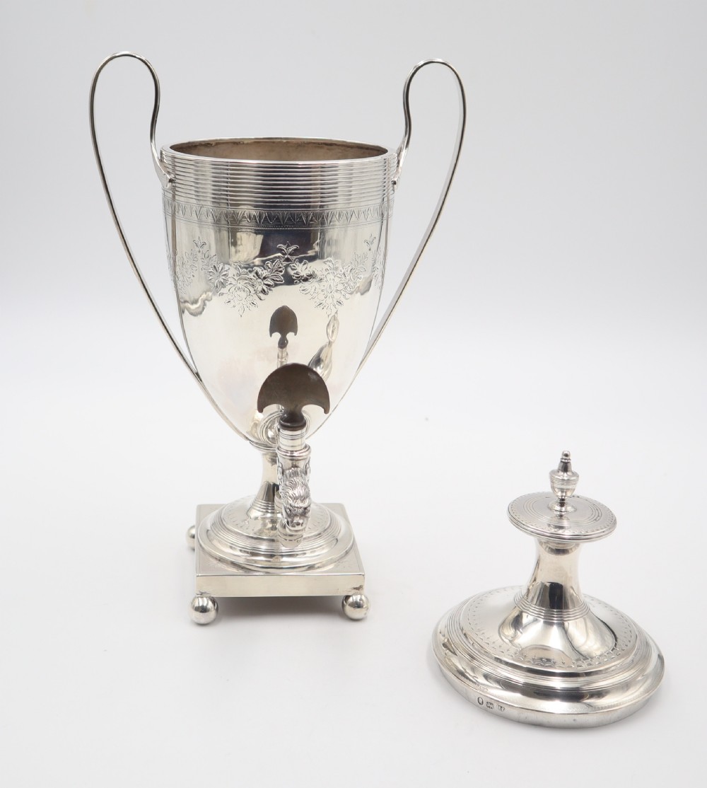 An antique silver tea urn featuring high loop handles, engraved floral bands, a front tap, and a square pedestal base supported by ball feet. View IX