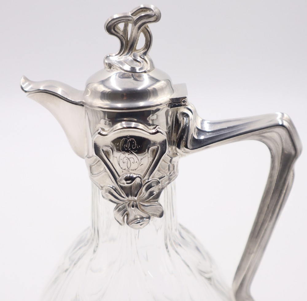 An Art Nouveau clear glass claret jug featuring a silver-plated collar, floral relief ornament, whiplash handle, and stylized lid finial. View VI