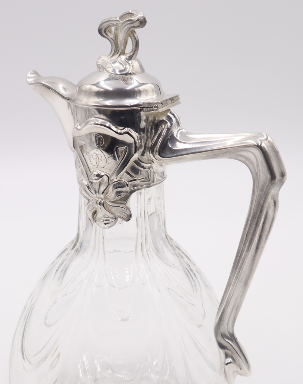 An Art Nouveau clear glass claret jug featuring a silver-plated collar, floral relief ornament, whiplash handle, and stylized lid finial. View VII