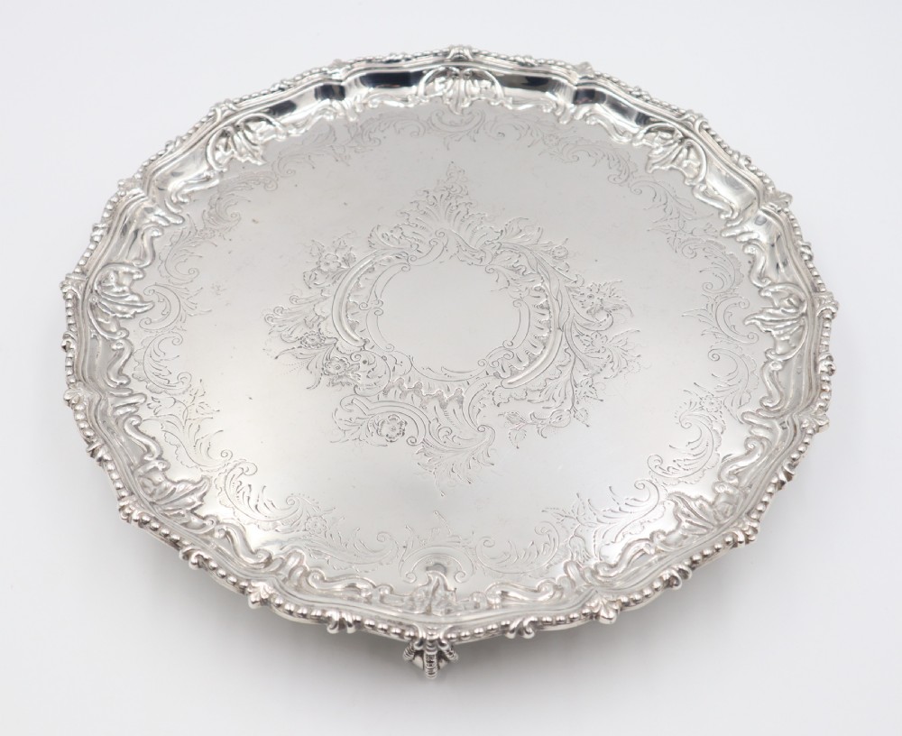 A circular silver salver featuring elaborate hand-engraved scrollwork, a raised shell border, and small cast feet.