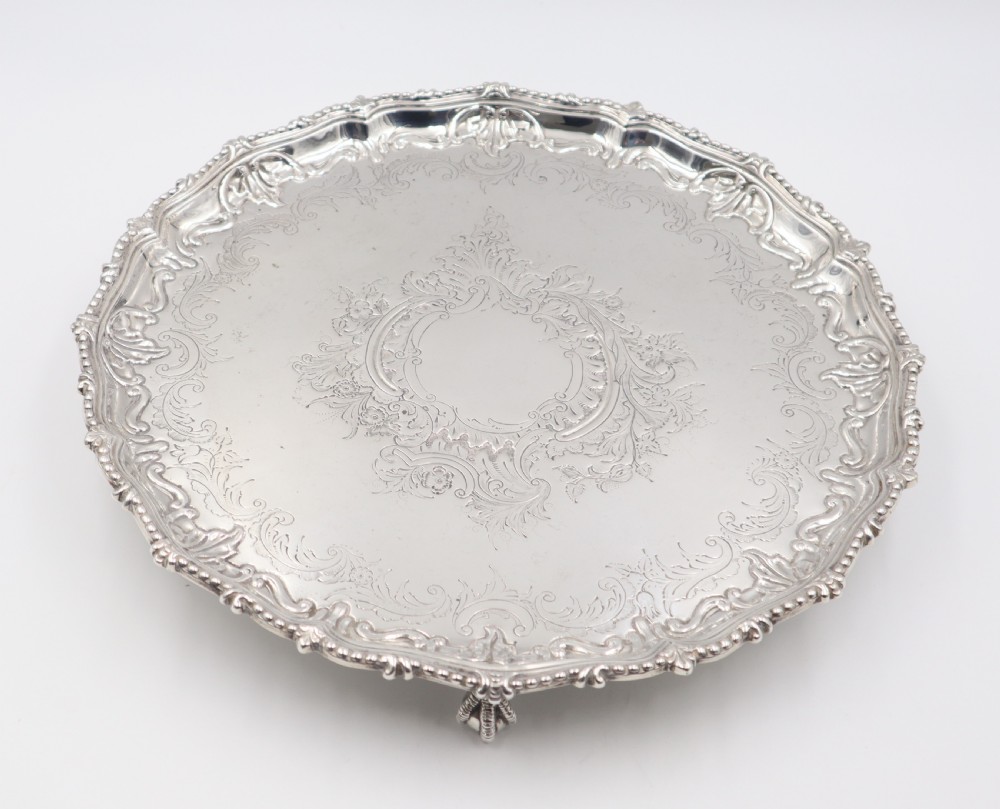 A circular silver salver featuring elaborate hand-engraved scrollwork, a raised shell border, and small cast feet. View II