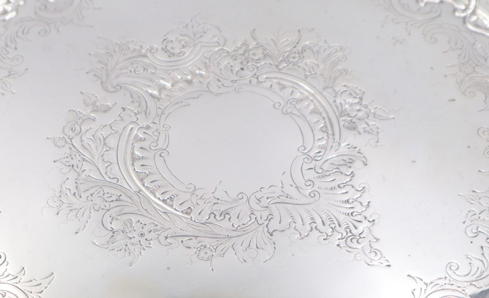 A circular silver salver featuring elaborate hand-engraved scrollwork, a raised shell border, and small cast feet. View III