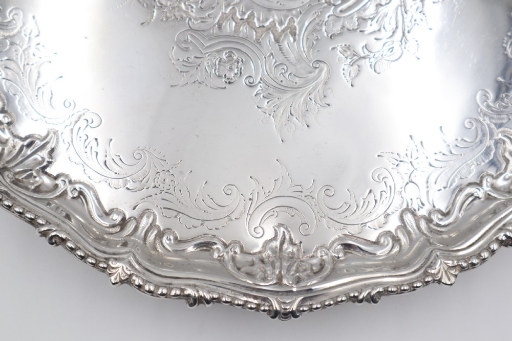 A circular silver salver featuring elaborate hand-engraved scrollwork, a raised shell border, and small cast feet. View IV