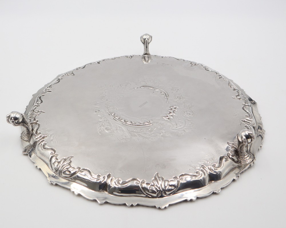 A circular silver salver featuring elaborate hand-engraved scrollwork, a raised shell border, and small cast feet. View VI